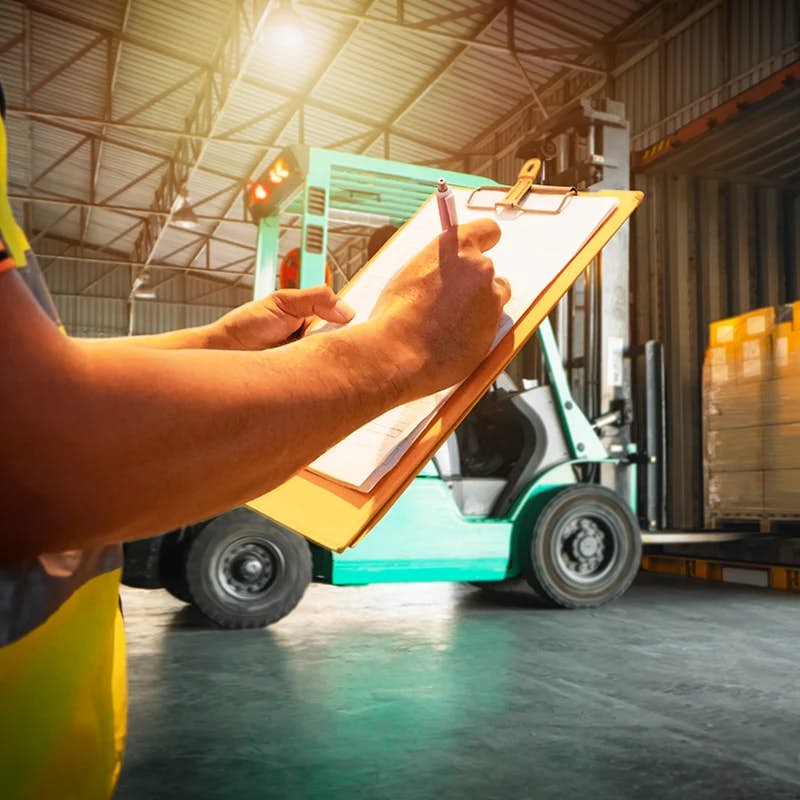 Close-up of a worker's hands holding a clipboard and pen, with notes or a checklist, in a warehouse setting. In the softly blurred background, a forklift is visible, suggesting active inventory management or preparation for loading goods.