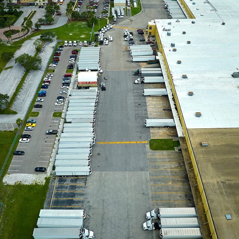 An aerial view of a busy distribution center with numerous trailers docked at loading bays. The parking lot is filled with cars, indicating a bustling logistics operation. The facility is flanked by greenery on one side, contrasting the industrial setting.