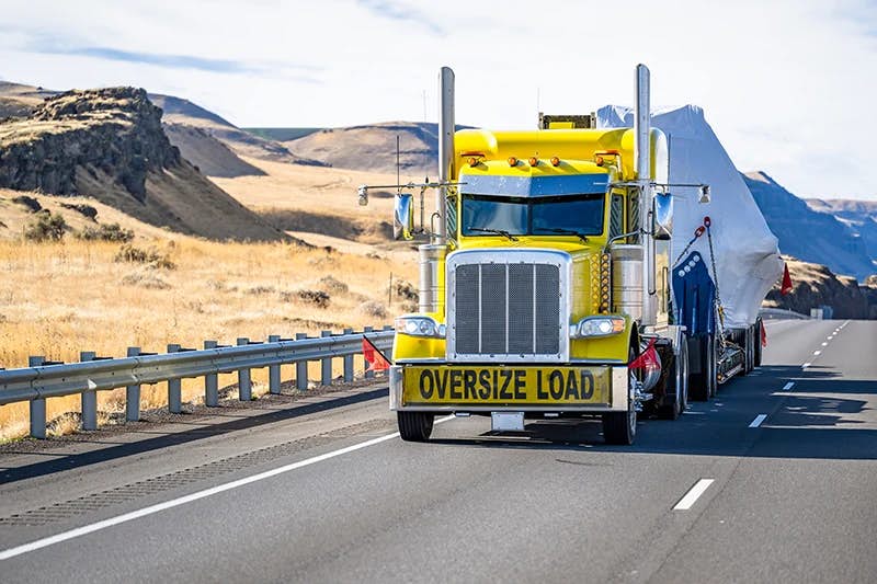 A striking yellow truck with chrome details is carrying an oversized load, as indicated by the banner on its front.