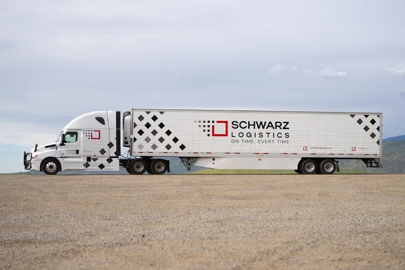 A large, modern white semi-truck with a long refrigerated trailer parked on a flat, expansive gravel surface.