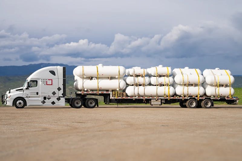 A modern white semi-truck connected to a step deck trailer, which is loaded with multiple large white tanks, each secured with yellow straps.