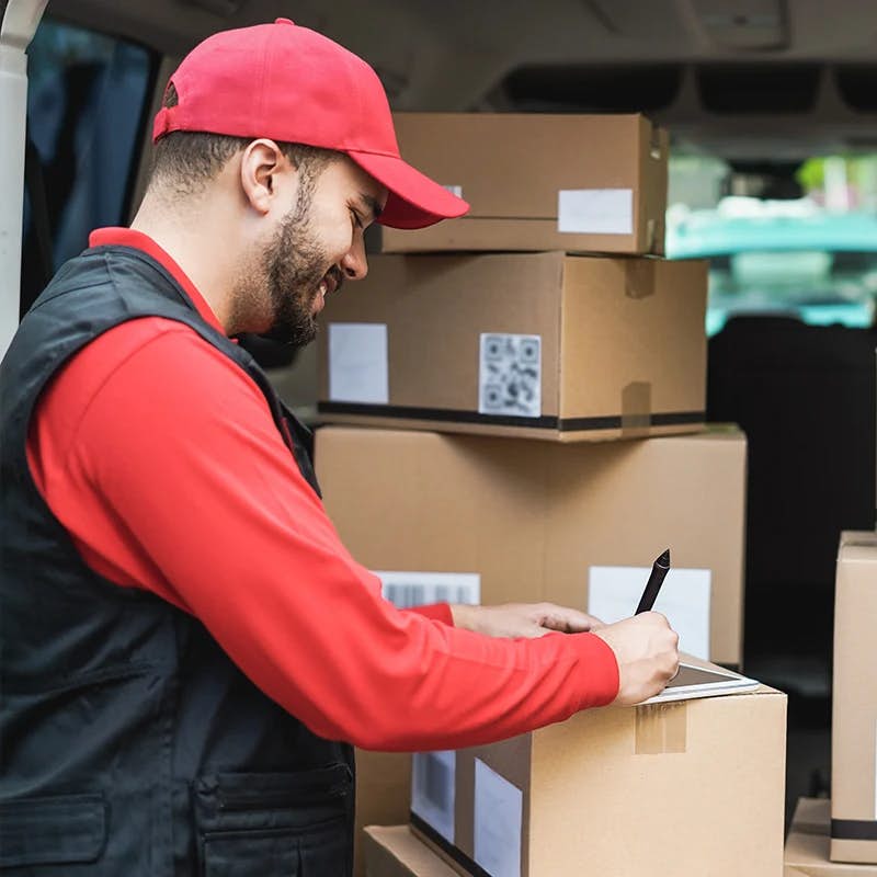 A delivery person in a red cap and a black vest with red sleeves, writing on a clipboard while leaning on a stack of cardboard boxes inside a delivery vehicle. The person is likely confirming details related to the parcels, such as tracking delivery status or checking inventory. QR codes visible on the boxes suggest that they are equipped for modern tracking methods.