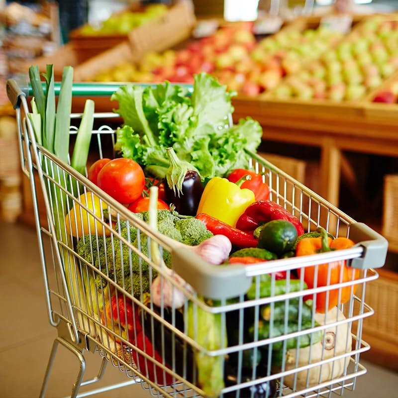 A shopping cart filled with an assortment of fresh vegetables, with a blurred background of a fruit stand.