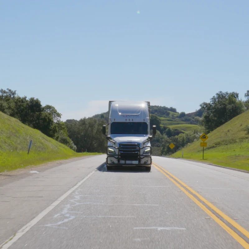A large semi-truck driving on a two-lane road. The setting is idyllic with green hills on either side under a clear blue sky, suggesting a peaceful, rural area.