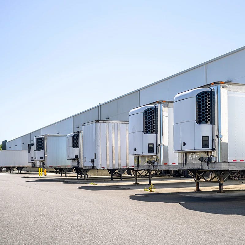Several white refrigerated trailers with vertical exhausts parked in a line, docked at a warehouse with a pale exterior under a clear blue sky.
