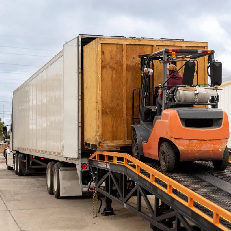 The image depicts a forklift loading a large wooden crate into the rear of a white dry van trailer. The forklift is orange and is driven by an individual who is partially visible. The trailer is parked, with its rear doors open, waiting to be loaded on an industrial or commercial lot. There's a portable, metal ramp extending from the ground to the trailer's deck, facilitating the loading process. The environment suggests an industrial or warehouse setting. The focus is on logistics and the movement of large
