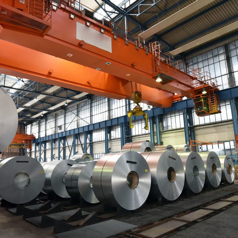 Large steel coils lined up in an industrial plant with an overhead crane system used for moving heavy materials.