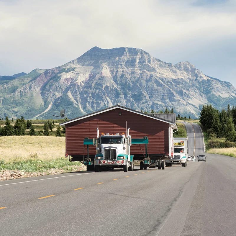 Truck hauling house on road with mountain backdrop.