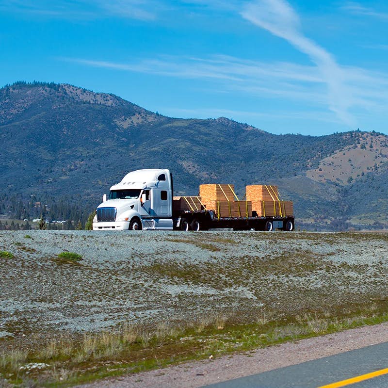 White semi truck hauling lumber on step deck trailer, mountains in background.