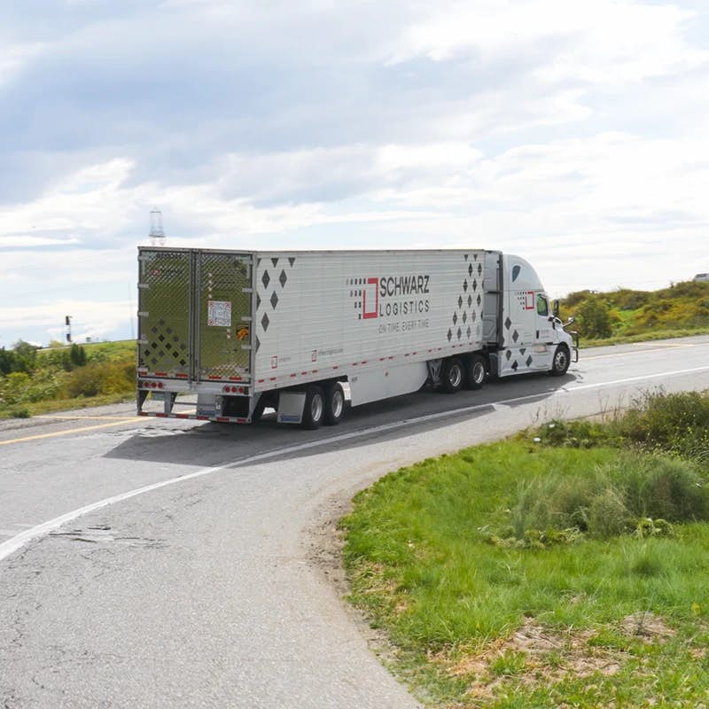 A white semi-truck with 'SCHWARZ LOGISTICS' branding is turning on a road, with its trailer doors open revealing a metal grate, against a rural backdrop with sparse greenery and a partly cloudy sky, suggesting a delivery or pickup in progress.
