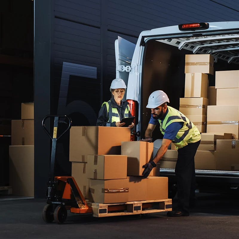 Two workers in safety vests and hard hats are busy with logistics work at night, with one holding a tablet and overseeing the loading process while the other manually moves boxes onto a pallet jack near an open delivery van, illuminated by the vehicle's interior light.