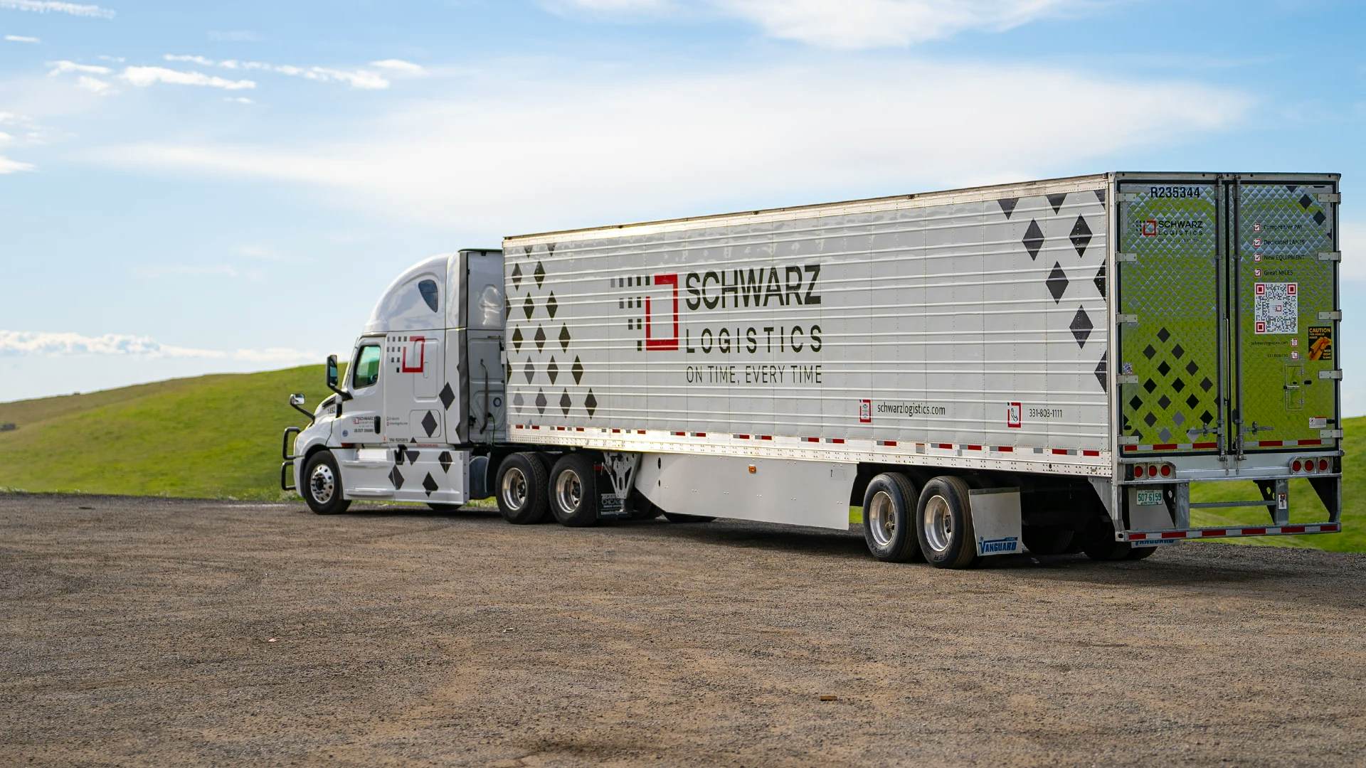 A white semi-truck with a trailer that has the name "SCHWARZ LOGISTICS" printed on the side, along with the tagline "ON TIME, EVERY TIME." The background shows a clear sky with lush green hills, and the truck is parked on a gravel lot.