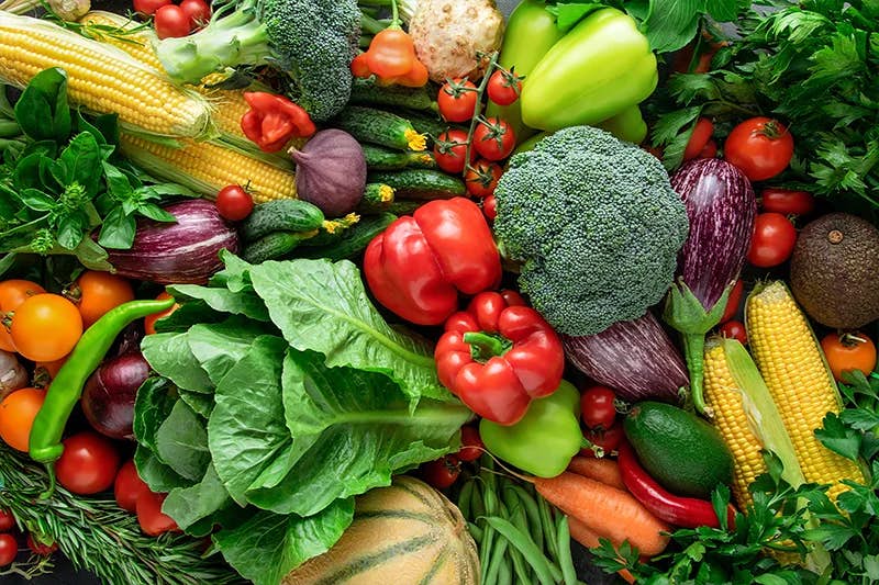A vibrant array of fresh vegetables and herbs densely packed together, featuring corn, broccoli, bell peppers, eggplants, carrots, tomatoes, and leafy greens, among others, offering a colorful feast for the eyes suggestive of healthy eating and agricultural bounty.