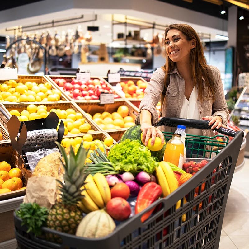 A smiling woman shopping in a grocery store, pushing a cart filled with a variety of fresh fruits and vegetables such as bananas, apples, and lettuce. She appears to be reaching for an item in the well-stocked produce section.