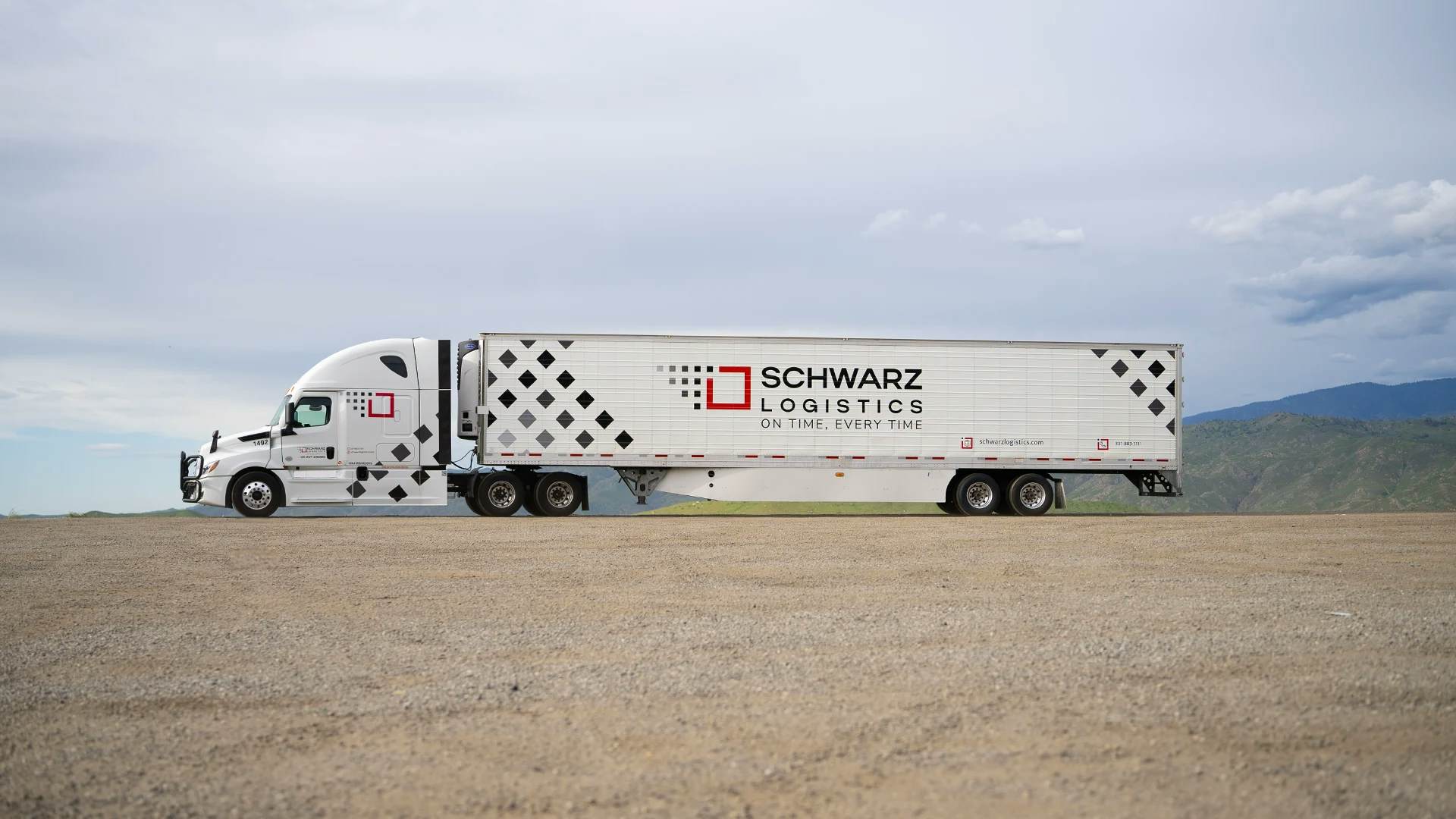 A white semi-truck with a large trailer parked on a flat, dry landscape. The trailer is branded with the name "SCHWARZ LOGISTICS" along with the slogan "ON TIME, EVERY TIME" and a geometric diamond pattern on its side. In the background, we see rolling hills under a cloudy sky, suggesting a vast, open rural or semi-arid environment. The truck appears stationary, likely captured in a moment between deliveries or while on a break during a long haul.