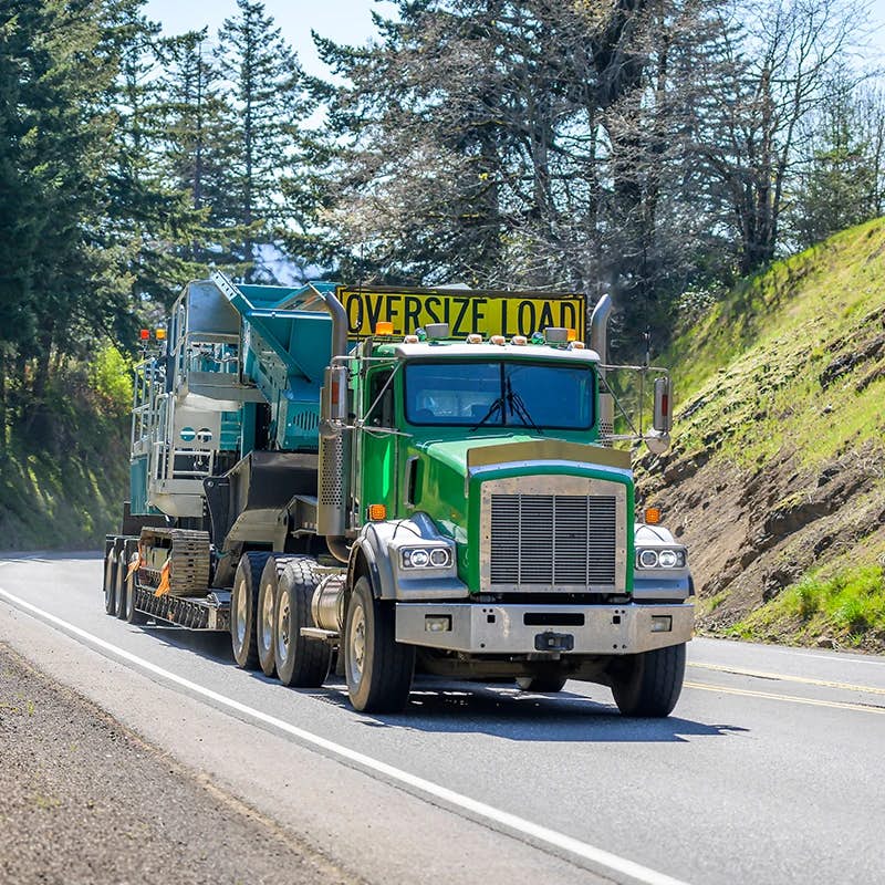 Green truck hauling oversized equipment on a sunny road with trees.