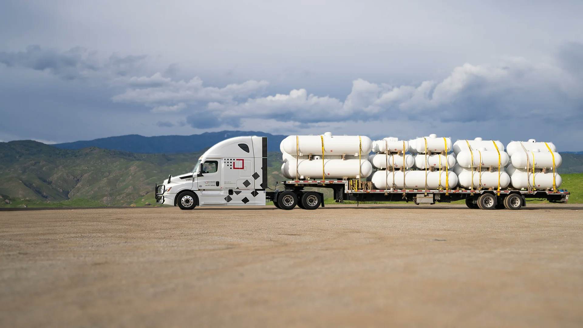 A white semi-truck hauling a flatbed trailer loaded with multiple large white industrial tanks, secured with yellow straps. The truck is parked on a concrete ground with a dramatic backdrop of dark storm clouds gathering over green hills, conveying a sense of impending weather challenge during transportation.