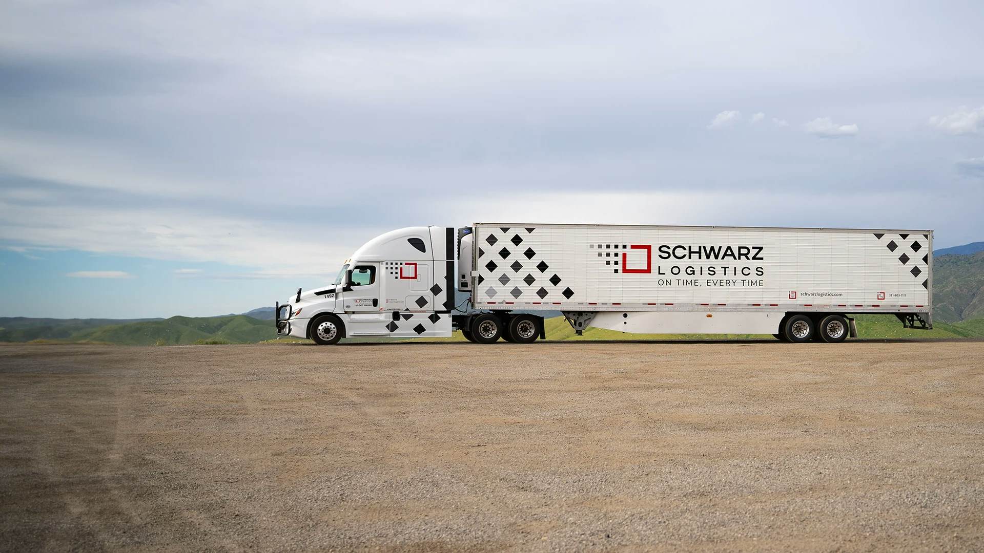A white semi-truck with a trailer, bearing the name "SCHWARZ LOGISTICS" and the slogan "ON TIME. EVERY TIME." The trailer's design includes a pattern of black and red diamonds, which makes the branding quite prominent.