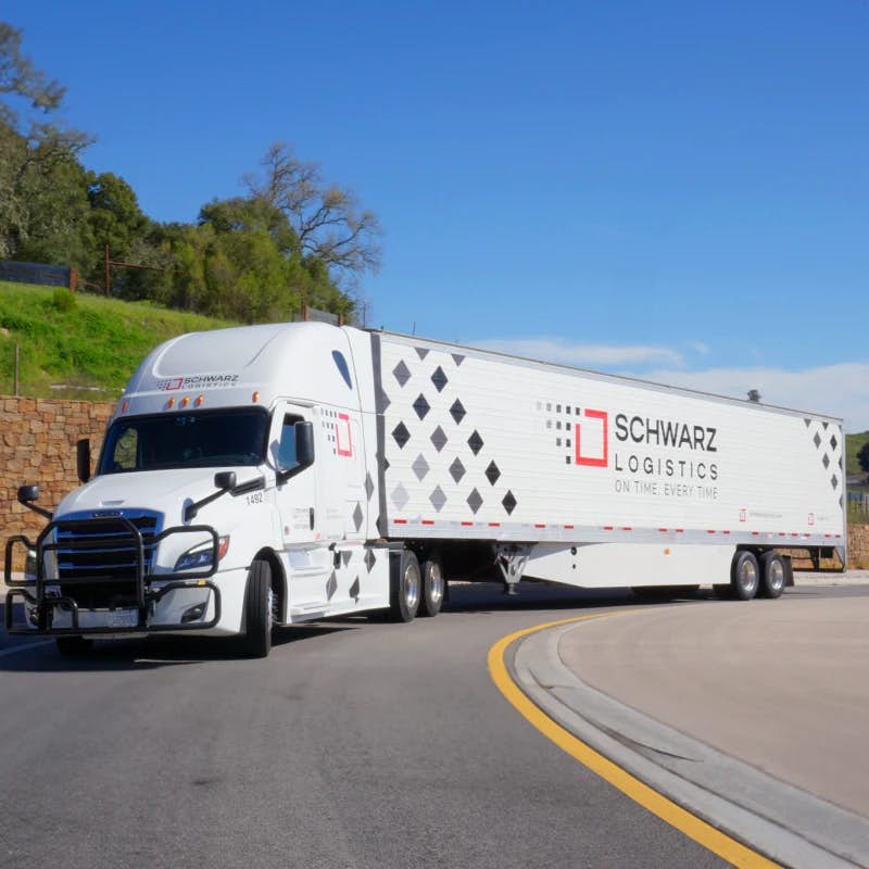 A white semi-truck with "SCHWARZ LOGISTICS" branding on the trailer is driving on the road, with a clear sky above and green trees in the background.