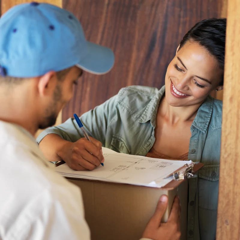 A delivery person in a blue cap is holding a clipboard and a pen, taking a signature from a smiling woman who is receiving a package. The image captures a friendly interaction, typically occurring at the customer's doorstep during a delivery service.