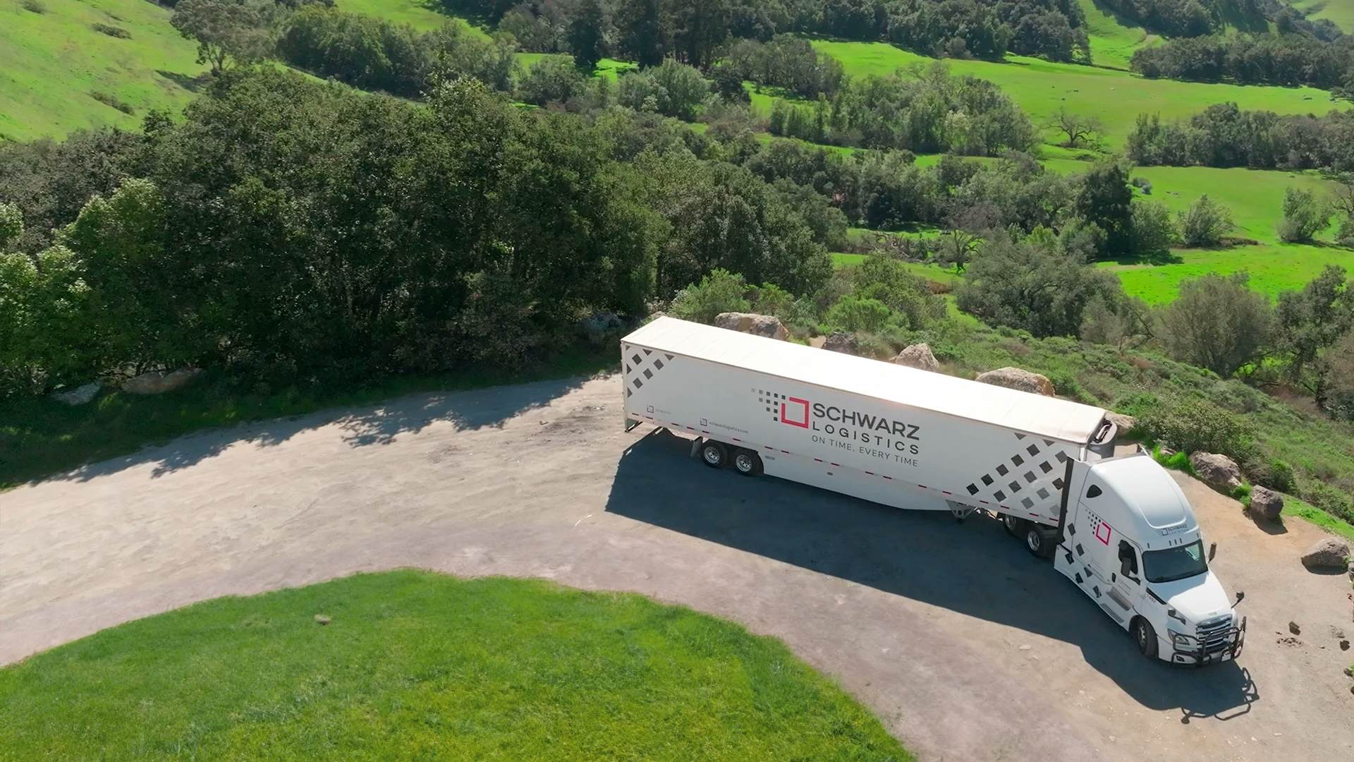Aerial view of a white semi-truck with "SCHWARZ LOGISTICS" branding on the trailer parked on a gravel lot, surrounded by lush greenery and hills.