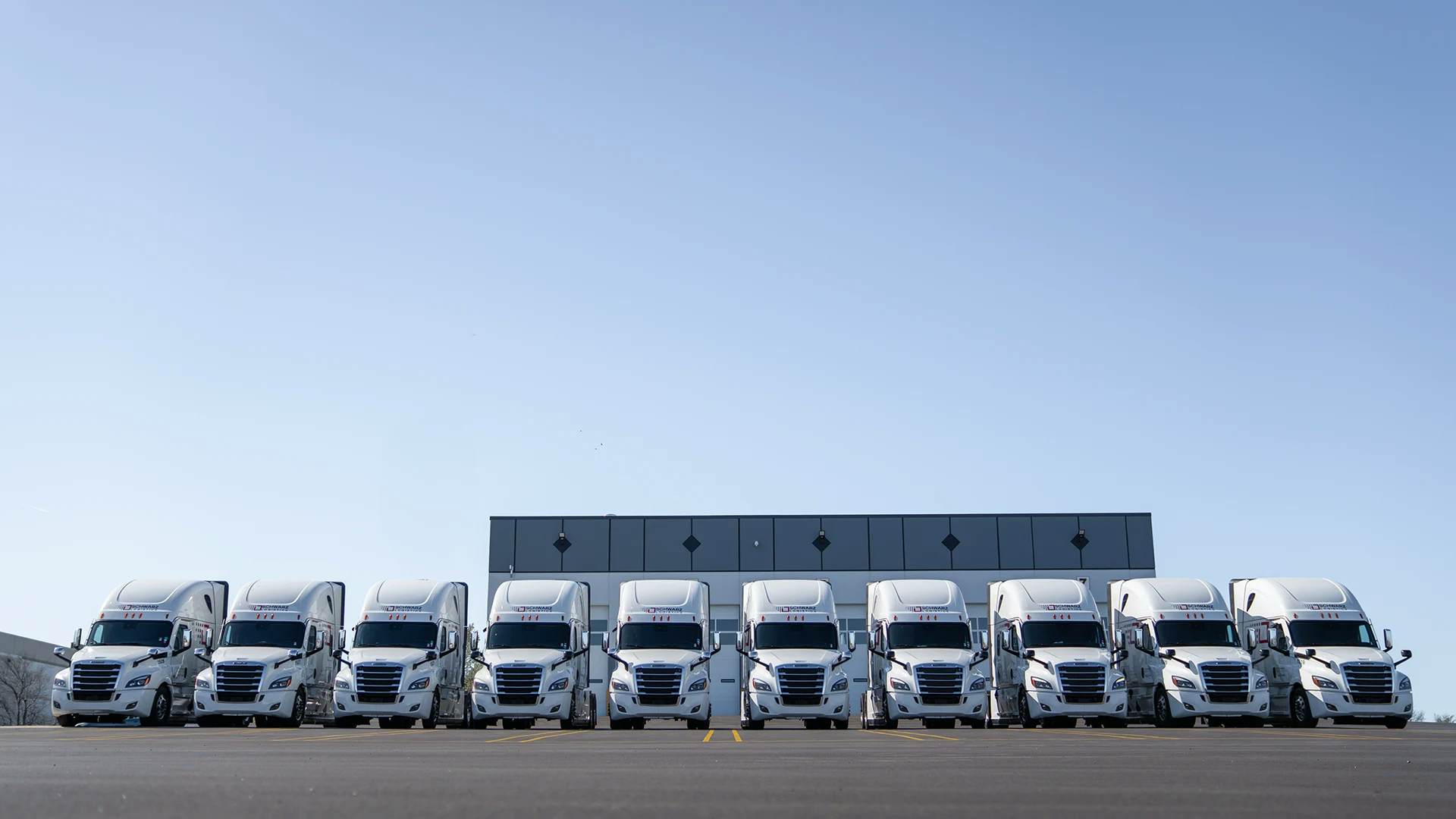 A fleet of white semi-trailer trucks arranged in a V-formation in a spacious parking area.