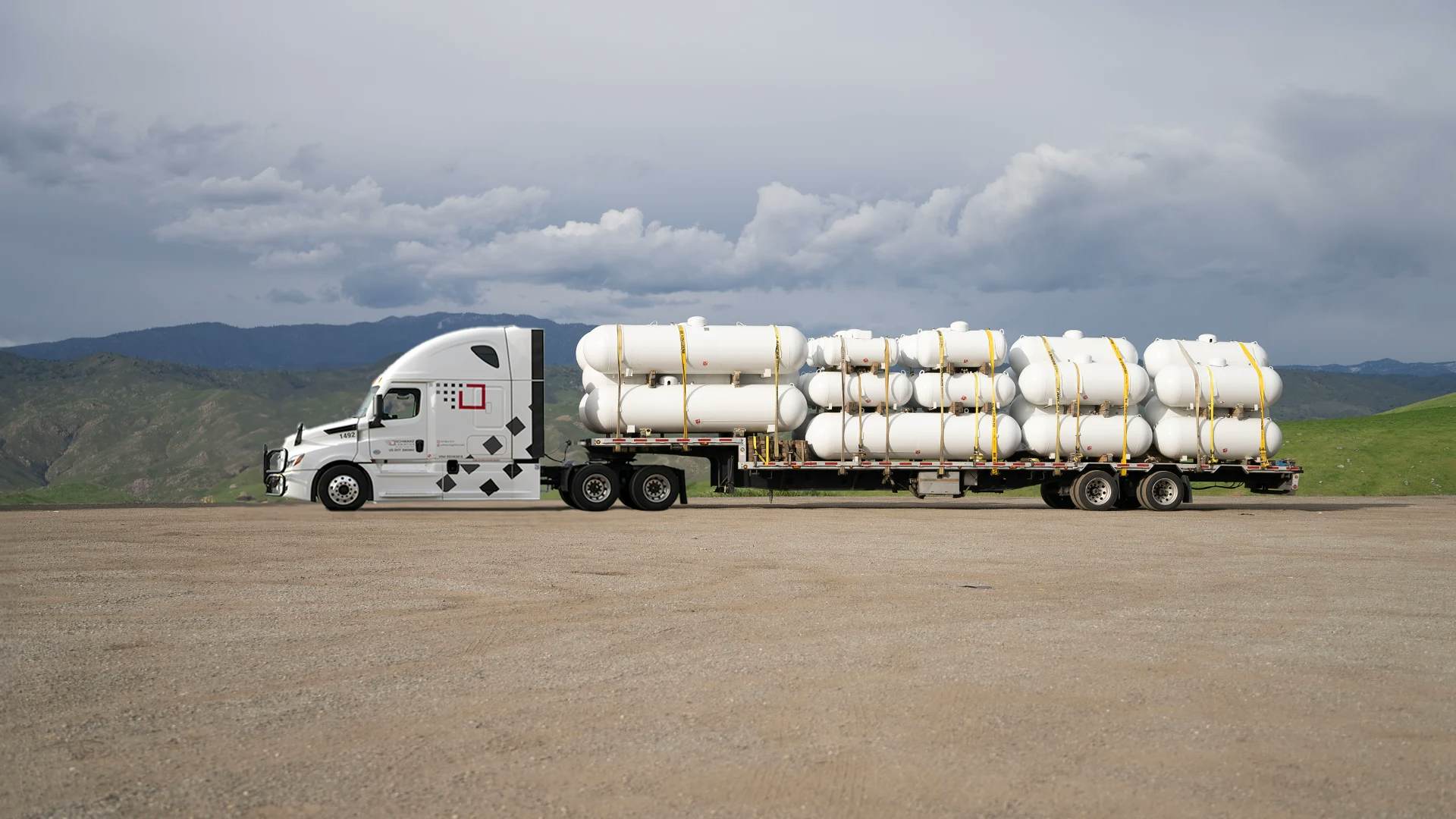 Semi truck hauling white tanks on step deck trailer, cloudy sky.