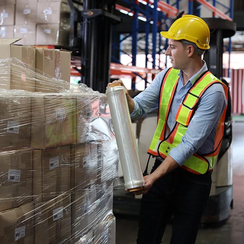 A warehouse worker in a safety helmet and high-visibility vest is using plastic wrap to secure a pallet loaded with boxes. The setting suggests a focus on safe and secure packaging practices in a distribution or storage facility.