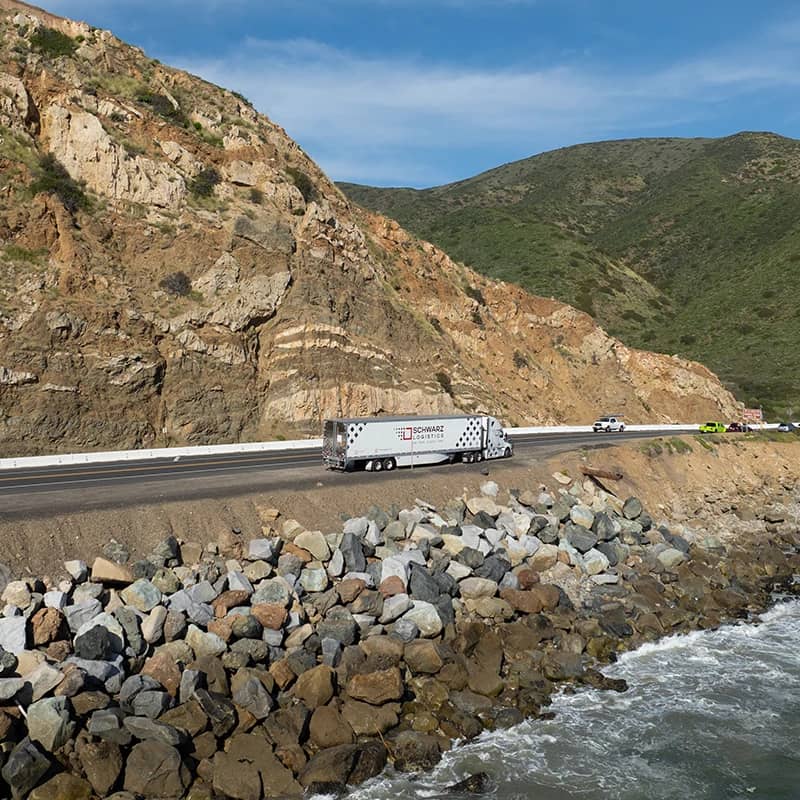 A "Schwarz Logistics" semi-trailer truck driving along a coastal highway with a steep cliff on one side and the ocean shore on the other.