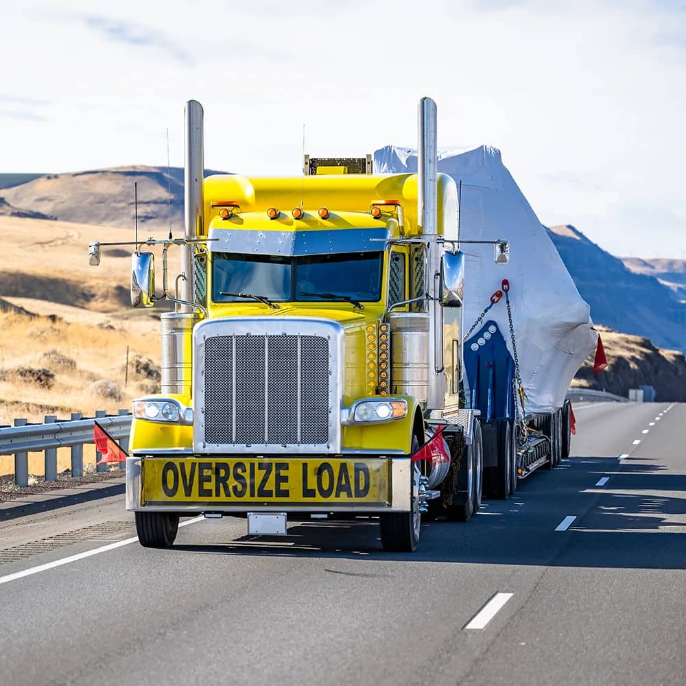 A close-up of enormous, stacked black tires secured on a flatbed trailer, with a prominent 'OVERSIZE LOAD' banner across the width and a red safety flag on the corner, indicative of the specialized transport of large industrial equipment.