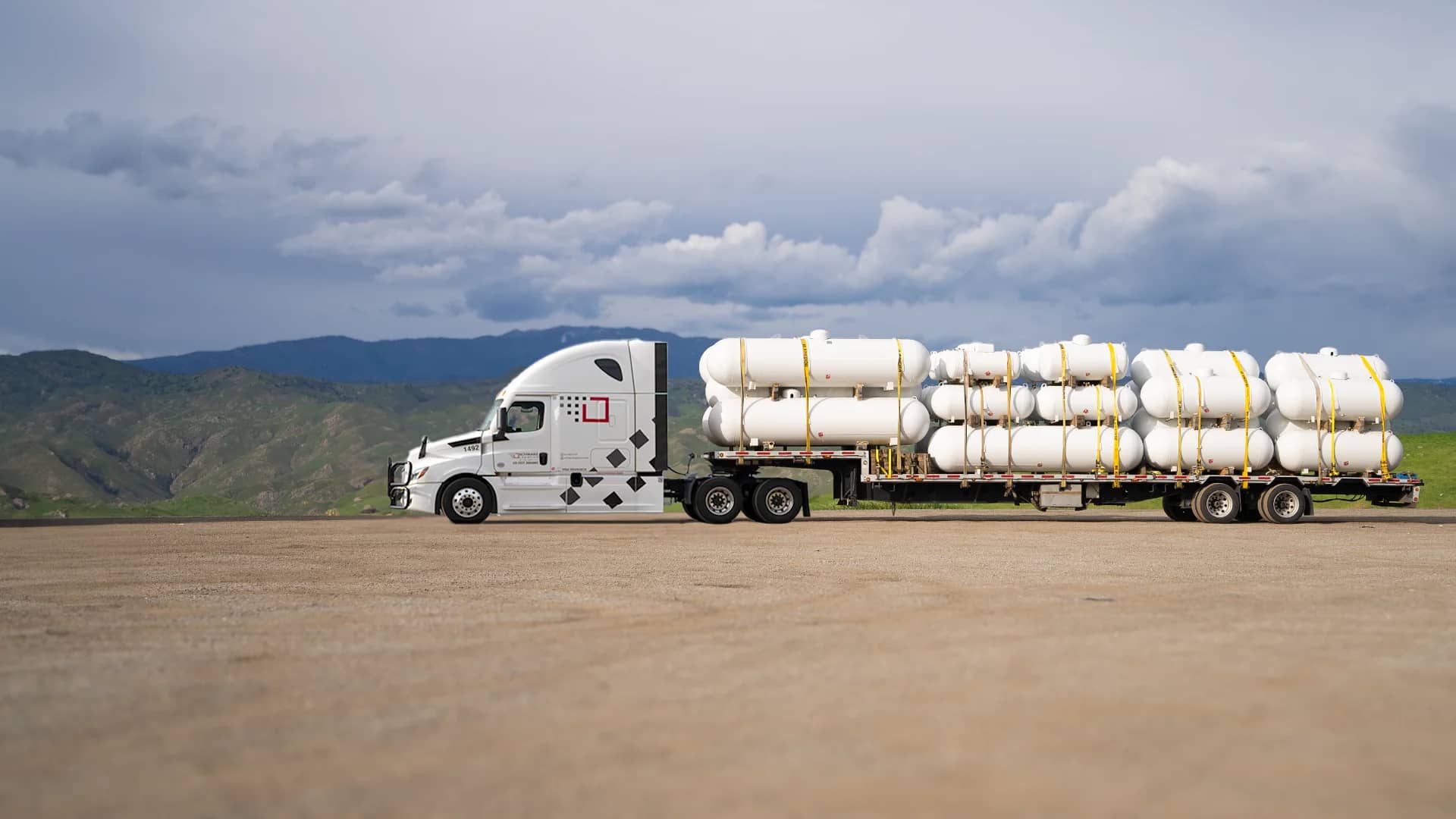 A white semi-truck hauling a flatbed trailer loaded with multiple large white industrial tanks, secured with yellow straps. The truck is parked on a concrete ground with a dramatic backdrop of dark storm clouds gathering over green hills, conveying a sense of impending weather challenge during transportation.